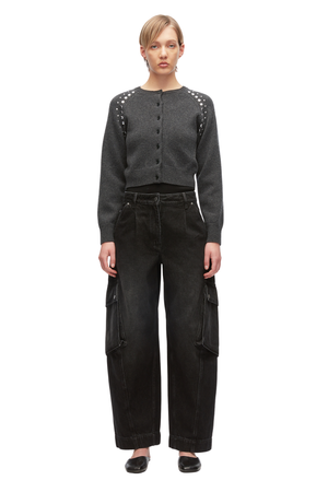 A model stands against a white background wearing a dark gray cropped button-front cardigan with silver stud embellishments on the shoulders, paired with black wide-leg cargo pants and black shoes.