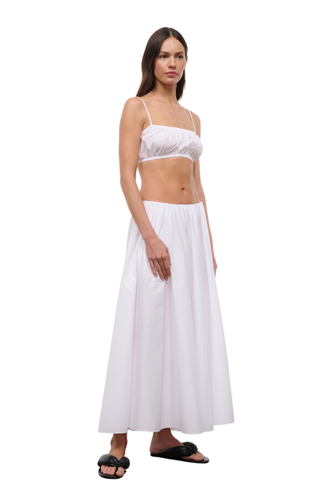 A woman modeling a white two-piece outfit consisting of a cropped camisole top with thin straps and a matching high-waisted maxi skirt, paired with black slide sandals, against a plain white background.
