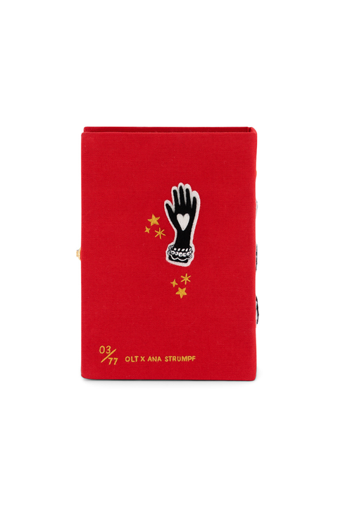 Back of a red book clutch featuring a black and white glove patch with a heart, yellow stars, and gold text reading '03/77 OLT X ANA STRUMPF' on a plain white background.
