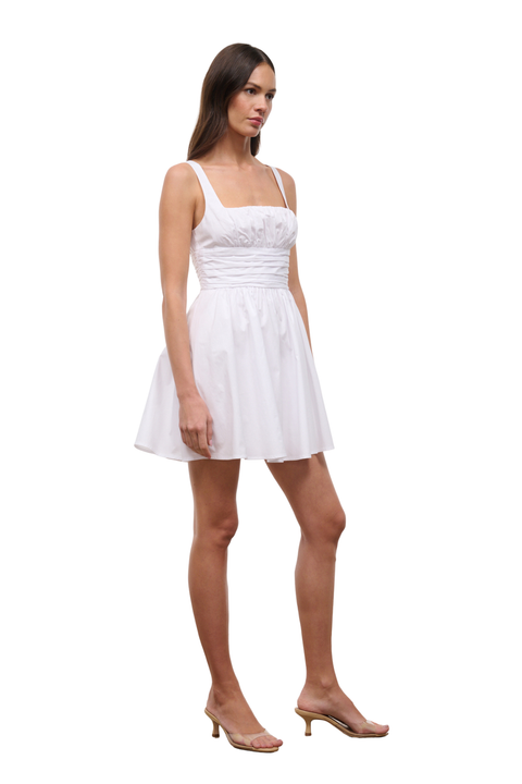 A woman modeling a short white sleeveless dress with a fitted bodice and flared skirt, paired with clear-strap heeled sandals, against a plain white background.