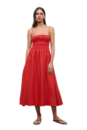 A woman modeling a bright red midi dress with thin spaghetti straps, a smocked bodice, and a flared skirt, paired with brown flip-flop sandals against a plain white background.