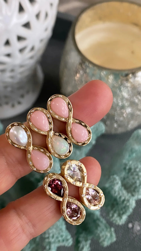 Woman displaying five five different infinity rings offered in different color combinations including, two pink opal, pink opal and white opal, pearl and pink opal, two red gemstones, and two clear gemstones.
