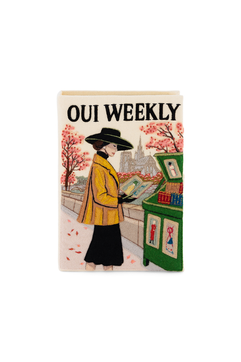 Book clutch with a vintage-style illustrated magazine cover titled 'OUI WEEKLY' on a plain white background. The cover shows a woman in a yellow coat and black hat standing beside a green newspaper stand, with pink trees and a cityscape in the background.