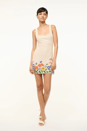 Woman wearing a cream glass-beaded, sleeveless mini dress that features a square neck, side slit, and intricate, colorful floral beading at the hem.