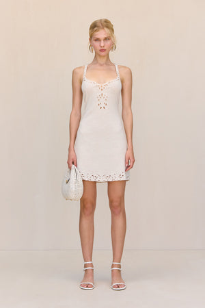 Woman wearing a cream knit mini dress that features an open floral embroidery along the neck like and the bottom hem. paired with a white handbag and white heels.