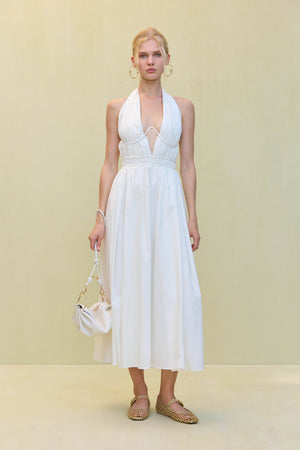 Woman wearing a white poplin midi dress that features a plunging halter neck and wire details at the bust, paired with a white handbag and tank sandals.