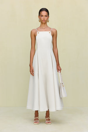 Woman wearing a white maxi dress that features a square neck with gold-toned grommet details attaching the spaghetti straps and a full skirt.