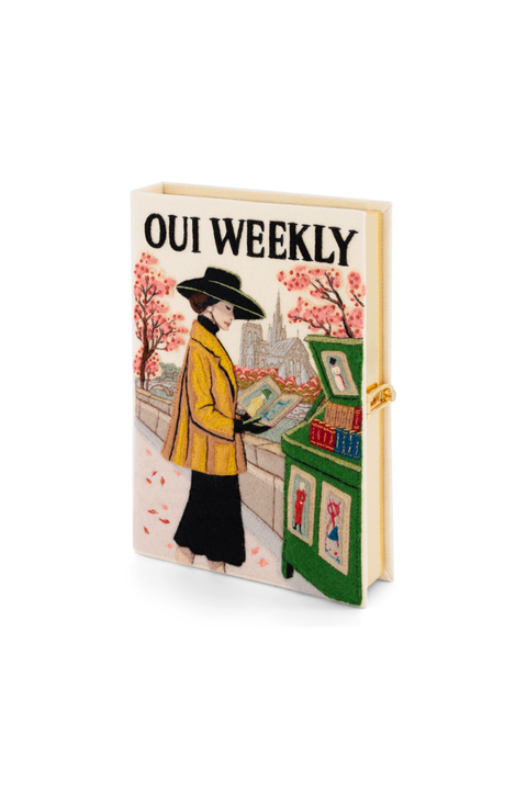 A book clutch titled 'OUI WEEKLY' featuring a vintage-style illustration of a woman in a yellow coat and black hat standing next to a green counter with pink cherry blossom trees in the background.