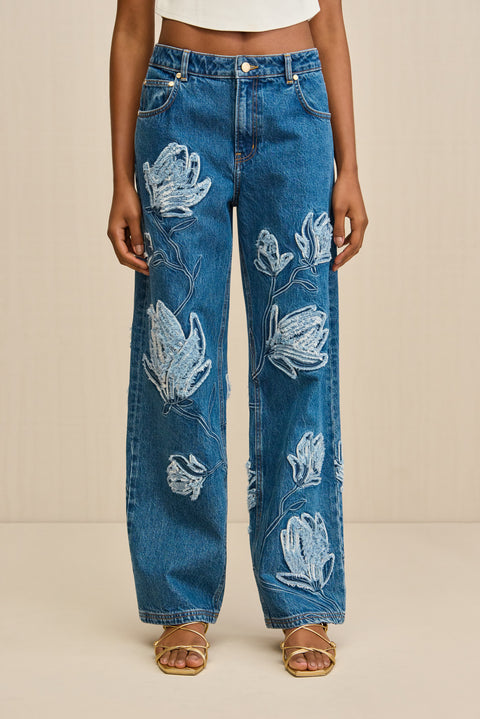 Close up of a woman wearing a pair of blue wash, wide leg jeans that feature an embroidered floral pattern in contrasting stitching.