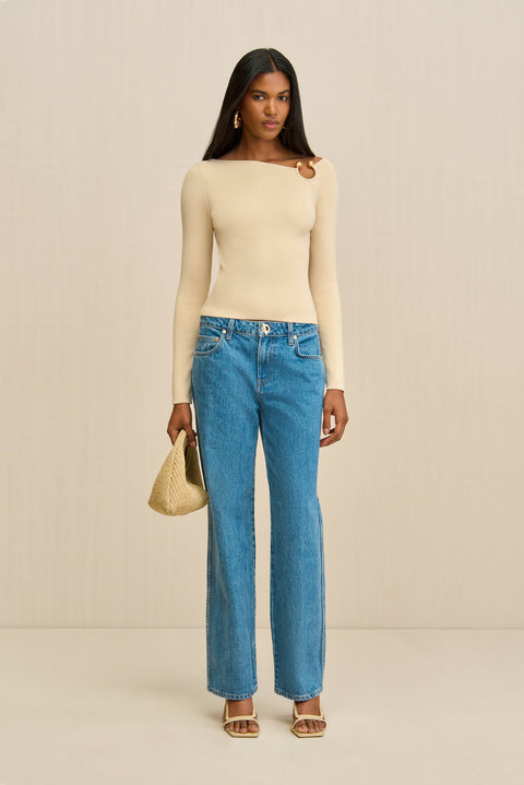 Woman wearing a cream long sleeve shirt with a ring detail at the shoulder, paired with blue jeans.