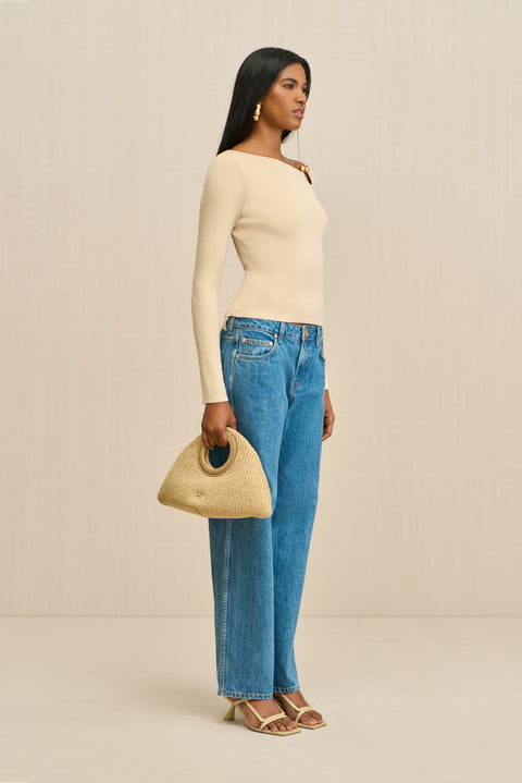 Side-facing woman wearing a cream long sleeve shirt with a ring detail at the shoulder, paired with blue jeans.