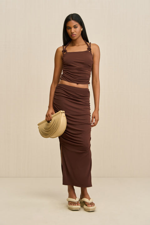 Woman wearing a brown tank top that features a knotted double strap detail and a matching brown maxi skirt, paired with tan sandals and handbag.