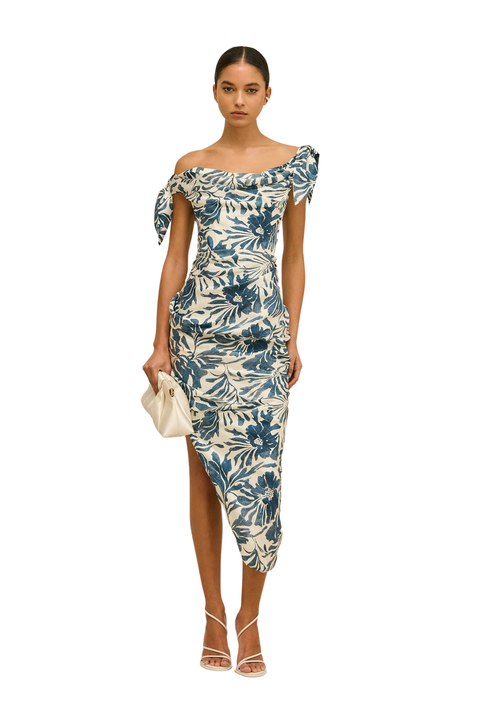 A woman modeling an off-the-shoulder midi dress with a blue and cream floral print, holding a small cream clutch bag against a plain white background.