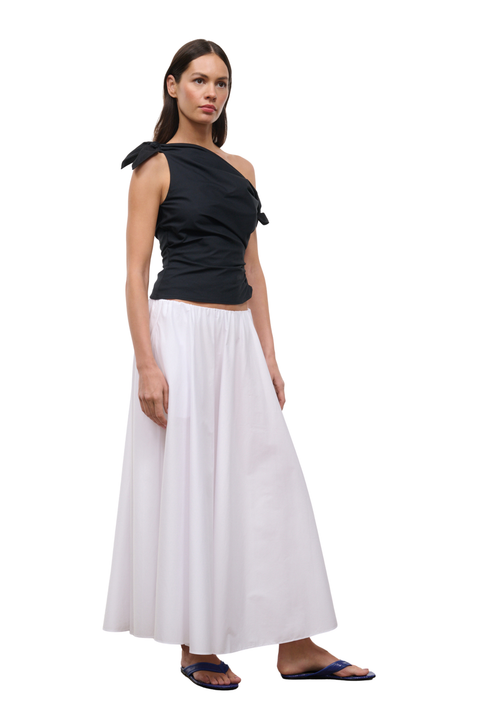 A woman modeling a two-piece outfit consisting of a black one-shoulder wrap top and a long white skirt, paired with dark blue flip-flop sandals, against a plain white background.