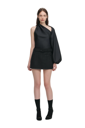 Woman wearing a black asymmetrical top and black mini skirt with belt with a white background