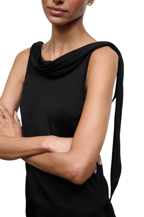 A close-up of a model wearing a black sleeveless top with a draped cowl neckline and a long tie detail on the shoulder, set against a plain white background.
