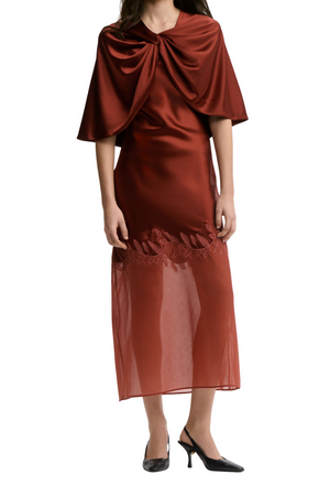 A model wearing a rust-brown satin dress with a draped, oversized shwal and a sheer skirt, styled with black heels against a plain white background.