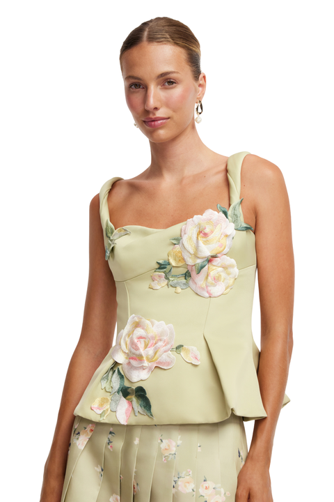 A woman modeling a light sage green sleeveless top with large 3D floral rose appliqués in pink, cream, and green, paired with a matching pleated skirt against a plain white background.
