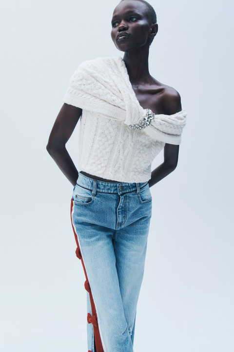 Woman wearing a white off-shoulder cable knit top and light blue jeans with red stripes on a plain background