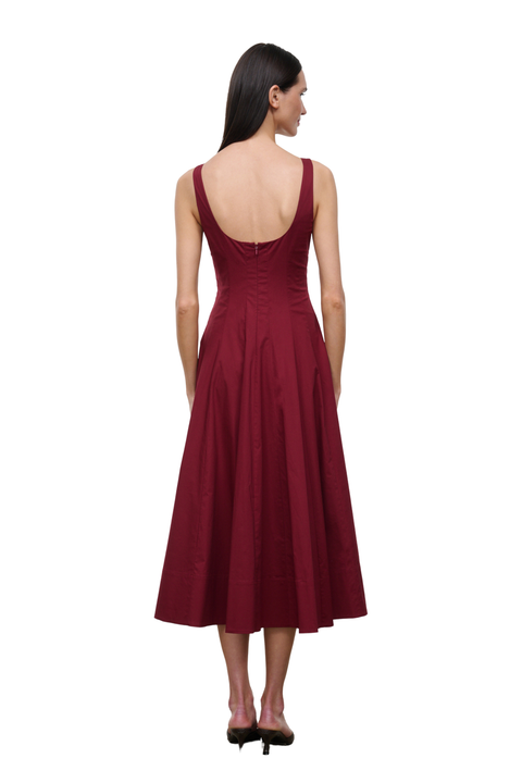 A woman viewed from the back wearing a sleeveless, deep burgundy midi dress with a fitted bodice, back tie detail, and flared skirt, paired with black heels against a plain white background.