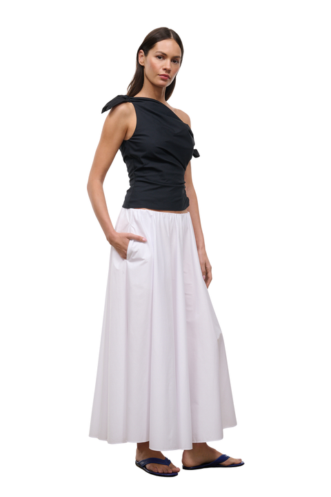 A woman modeling a black one-shoulder wrap top and a long white skirt with side pockets, paired with dark blue flip-flop sandals, against a plain white background.
