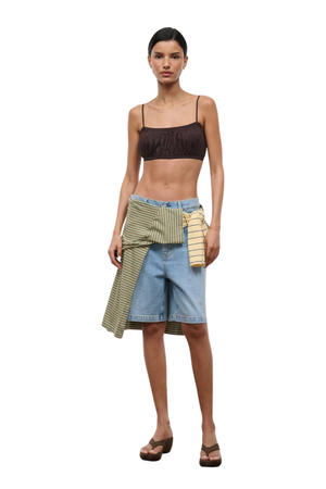 A model stands against a plain white background wearing a dark brown ribbed crop top, light blue denim shorts, and a striped green sarong tied around her waist with a matching striped pouch.