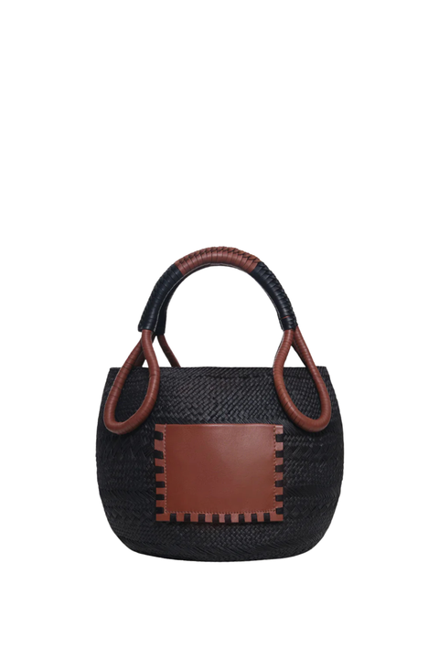 A small woven black straw handbag with brown leather trim, featuring a rectangular brown leather front patch and a curved brown leather top handle, shown against a plain white background.