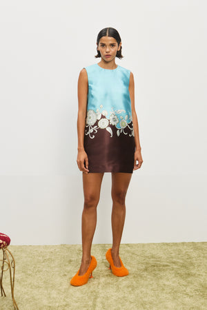 Woman wearing sleeveless, mini shift dress that is turquoise on the top and brown on the bottom, with a white floral pattern merging the two colors at the waist, paired with orange heels.