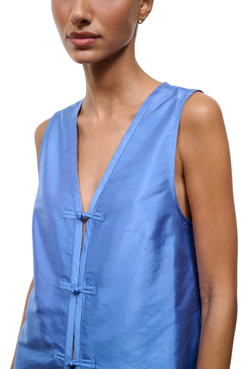 A close-up of a model wearing a sleeveless, V-neck top in a medium blue fabric with tie closures down the front.