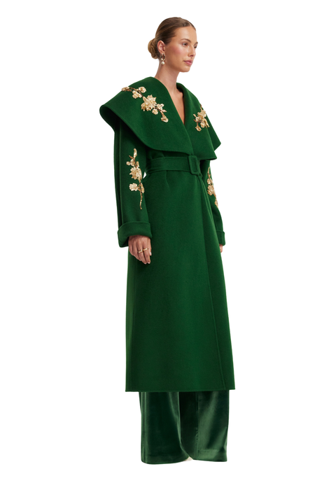 A model is shown from the side wearing a dark green wool coat with gold floral embroidery on the shoulders and sleeves, paired with matching green velvet trousers, against a plain white background.
