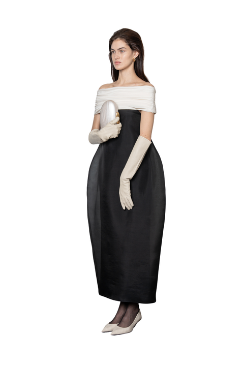 A woman modeling a black off-the-shoulder maxi dress with a white off-the-shoulder top band, long cream gloves, and pointed-toe heels against a plain white background.