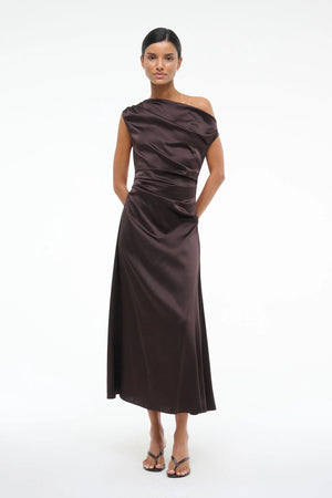 Woman wearing an asymmetrical, brown silk midi dress that features an off the shoulder design with draped ruching throughout, paired with brown sandals.