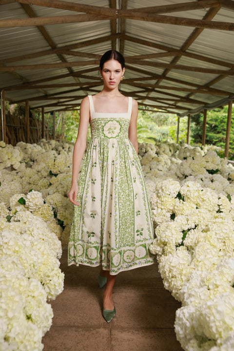 Model wears floral print midi dress next to rows of white flowers.