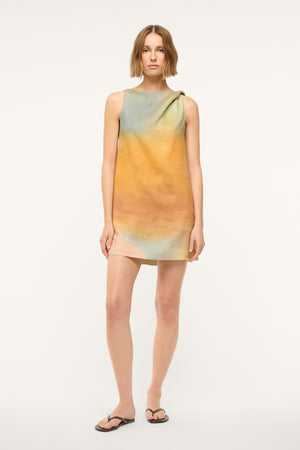 Woman wearing a sleeveless, pastel multicolor, mini shift dress that features a twist detail on one shoulder, worn with black sandals.