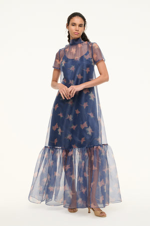 Woman wearing an organza gown in a navy floral print that features a mock neck, tiered ruffle hem, and removable slip, paired with tan heels.