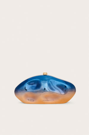 An abstract oval shaped acrylic clutch that features formed dents and a metallic blue to orange ombre effect, finished with gold hardware.
