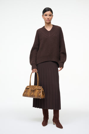 Woman wearing a brown ribbed sweater and skirt holding a patterned handbag on a white background