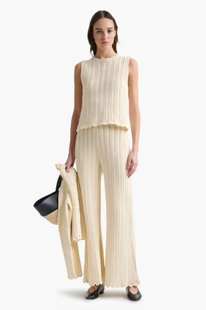 Woman wears knitted cotton tank top with ripple stitch and matching pants holding sweater and handbag.