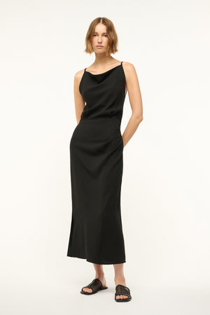 Woman wearing a black midi-length column dress that features a cowl neck, spaghetti straps, and a side slit, paired with black sandals.