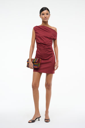 Woman wearing a deep red mini dress that features an asymmetrical off-the-shoulder neckline and draped ruching throughout, paired with black kitten heels.