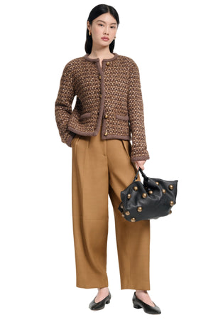 Woman wearing a brown tweed jacket and tan pants, holding a black handbag on a white background