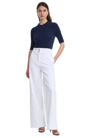 Woman wearing a navy top and white straight leg pants with gold buttons on a white background