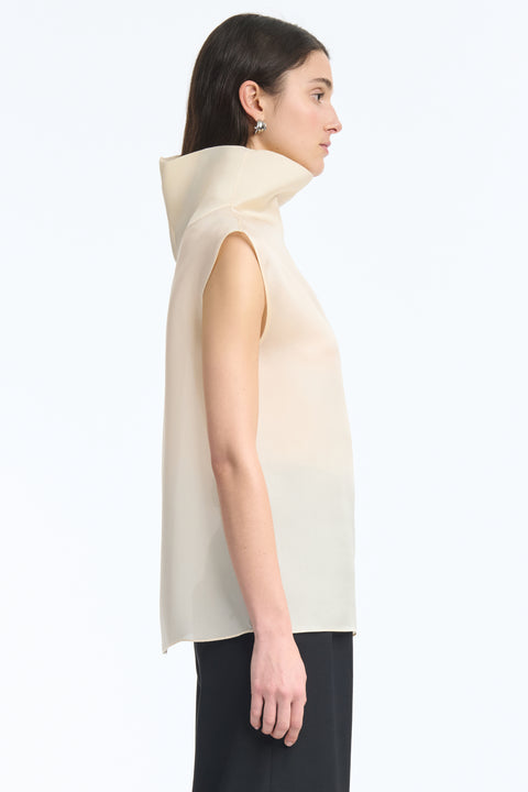 Side view of woman wearing a translucent high-neck sleeveless cream top and black pants on a white background