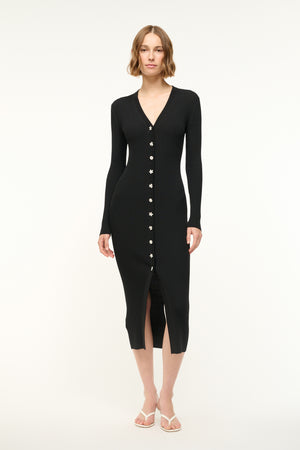 Woman wearing a black ribbed-knit, long sleeve midi dress that features silver toned floral buttons down the front, paired with white sandals.