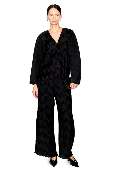 A model wearing a black textured long-sleeve wrap top and matching wide-leg trousers with a subtle geometric pattern, styled with black pointed flats and a small black clutch against a plain white background.