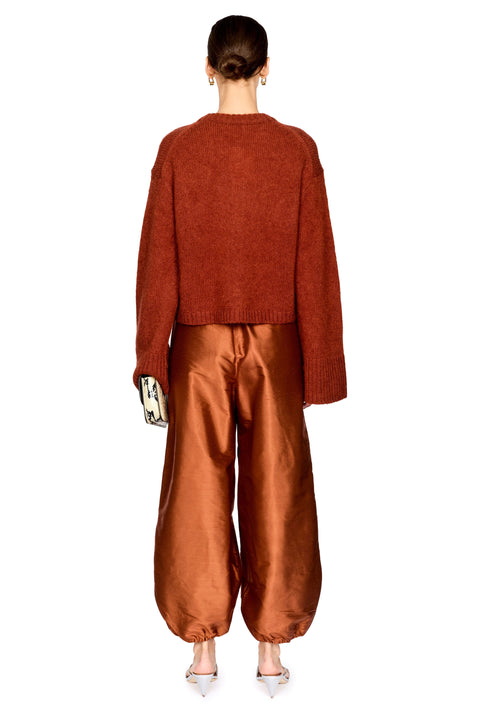 A model shown from the back wearing a textured rust-orange knit sweater and shiny bronze-brown wide-leg pants, paired with silver shoes against a plain white background.