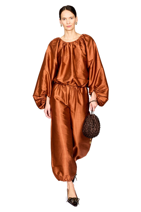 A model wearing a matching rust-brown satin two-piece set with a gathered, balloon-style top and wide-leg pants, holding a small woven brown handbag against a plain white background.