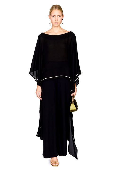 A woman modeling a black pants and a matching top with wide draped sleeves and thin white piping accents, holding a small green handbag against a plain white background.