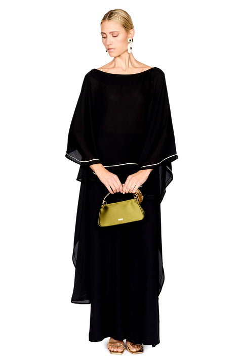 A woman modeling a black pants and a matching top with wide draped sleeves and thin white piping accents, holding a small green handbag against a plain white background.