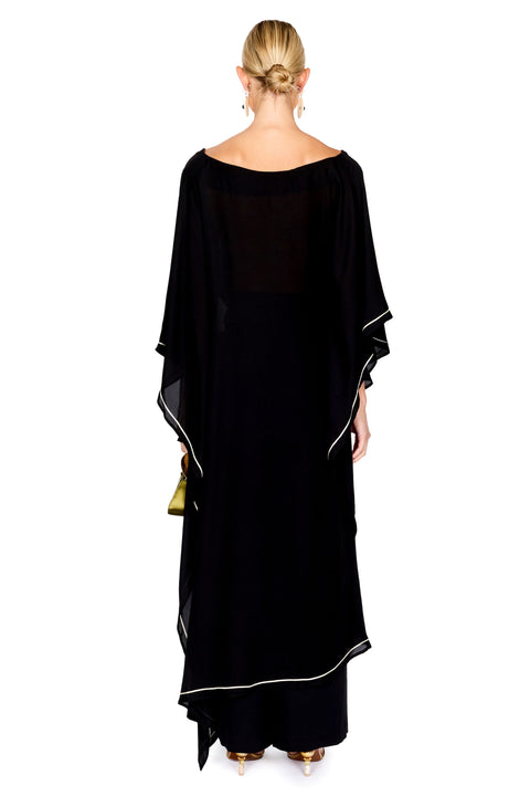 A woman modeling a black pants and a matching top with wide draped sleeves and thin white piping accents, holding a small green handbag against a plain white background.